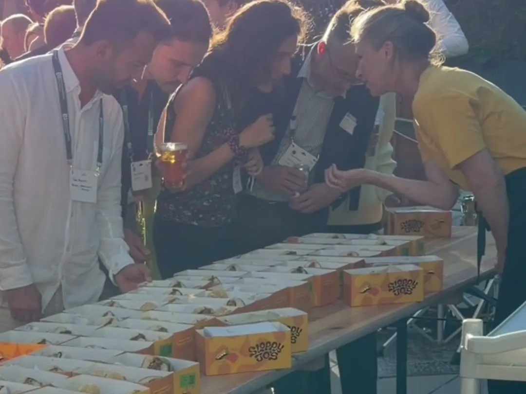 Guests at BioInnovation Institute Demo Day lifting Sloppy Jo's boxes from a long table in evening sunlight, while a Sloppy Jo's team member serves