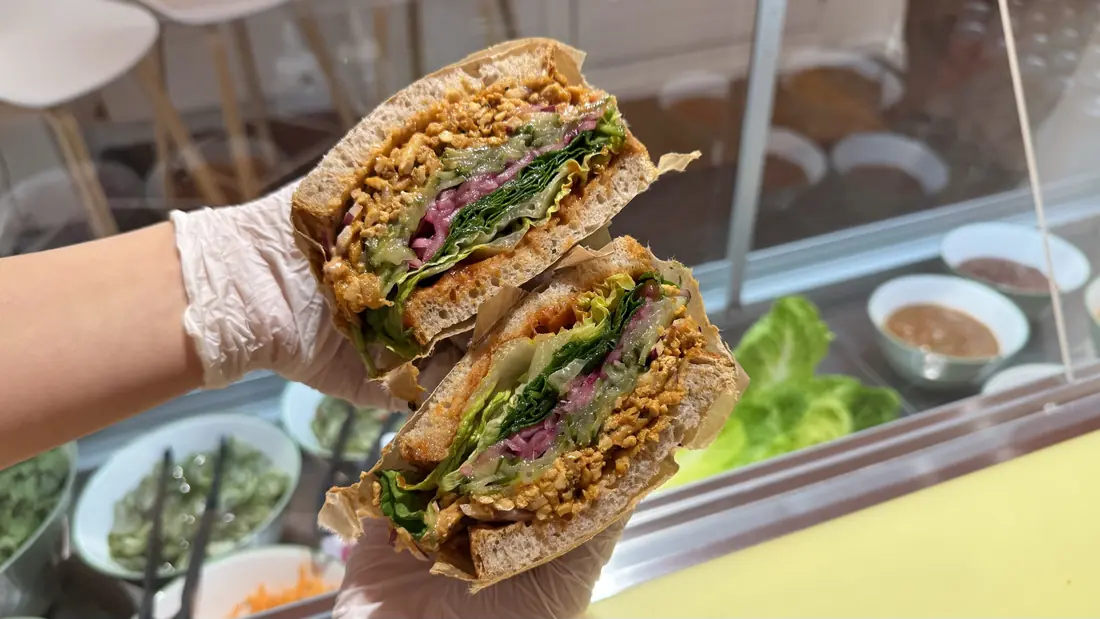 Sloppy Jo's plant-based deli sandwich on Meyers Bageri sourdough, cut in half and held up over the deli's prep counter - shredded tofu filling, pickled red onion, fresh lettuce and chili sauce visible, with fresh ingredients in the background