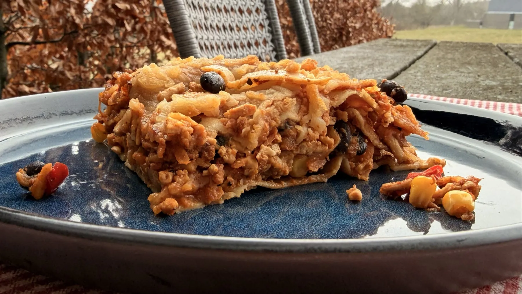 A slice of Mexican lasagne on a deep blue plate, layers of pasta, beans, corn, and golden-topped filling clearly visible